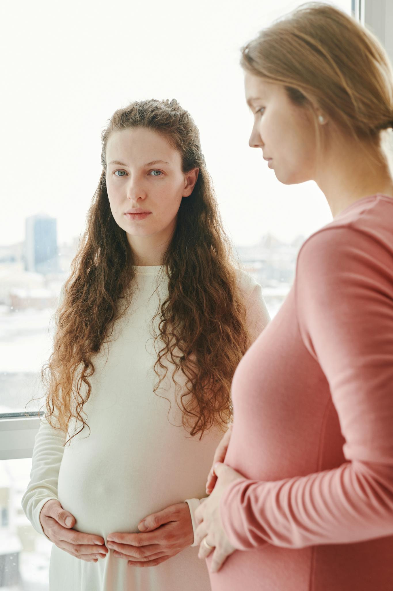 Two pregnant women standing by a window, hands on bellies, embracing motherhood.